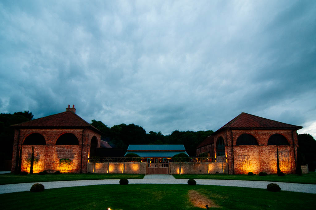 A beautifully lit up Hazel Gap Barn, surrounded by lush green trees and tranquil countryside. The stone walls of the barn are illuminated, casting a warm and inviting glow, creating a romantic and serene atmosphere for weddings and events. The lights highlight the charming, rustic architecture of the barn and create a stunning visual contrast against the night sky.