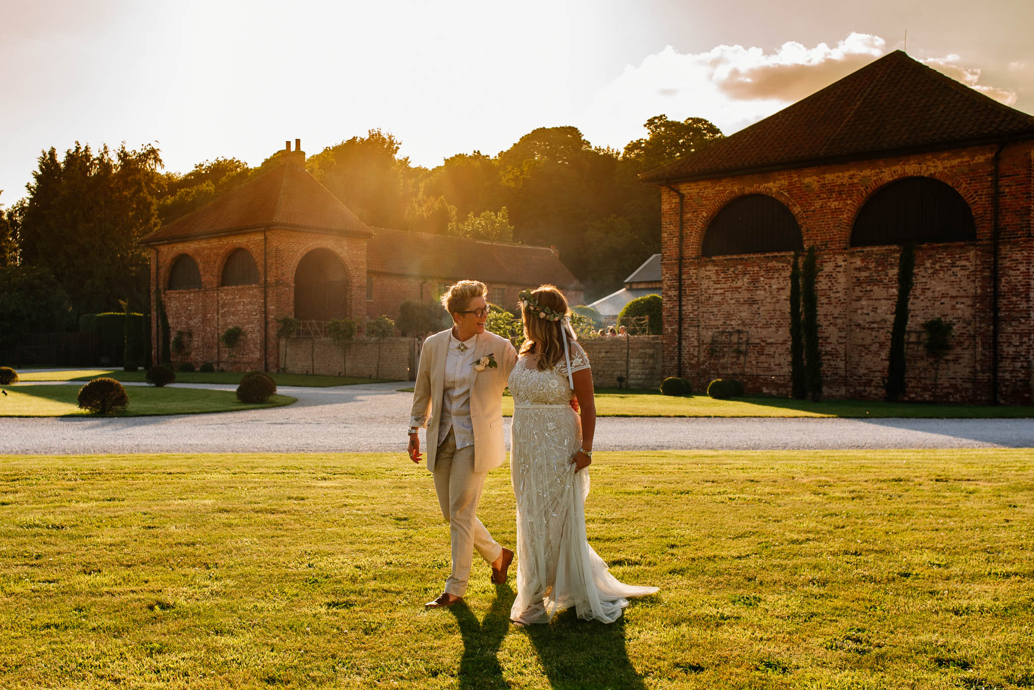 Two brides walking hand in hand, pictured against a stunning sunset with Hazel Gap Barn in the background. The sun is setting behind the venue, casting a warm orange and pink glow over the countryside. Hazel Gap Barn offers a breathtaking setting for a unique and intimate wedding celebration, surrounded by nature and breathtaking scenery.